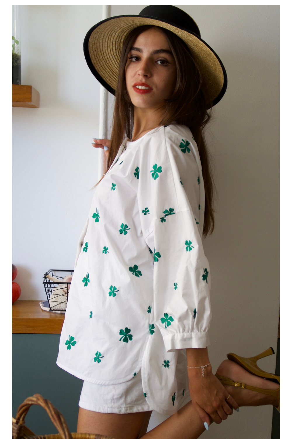 Femme brune de profil portant la blouse Dublin de Carlis, blanche avec trèfles verts brodés, manches ballons et dos plus long. Elle porte un chapeau de paille et lève une jambe pour montrer un talon. Photographie de style éditorial dans une cuisine pour cette création faite main en France.