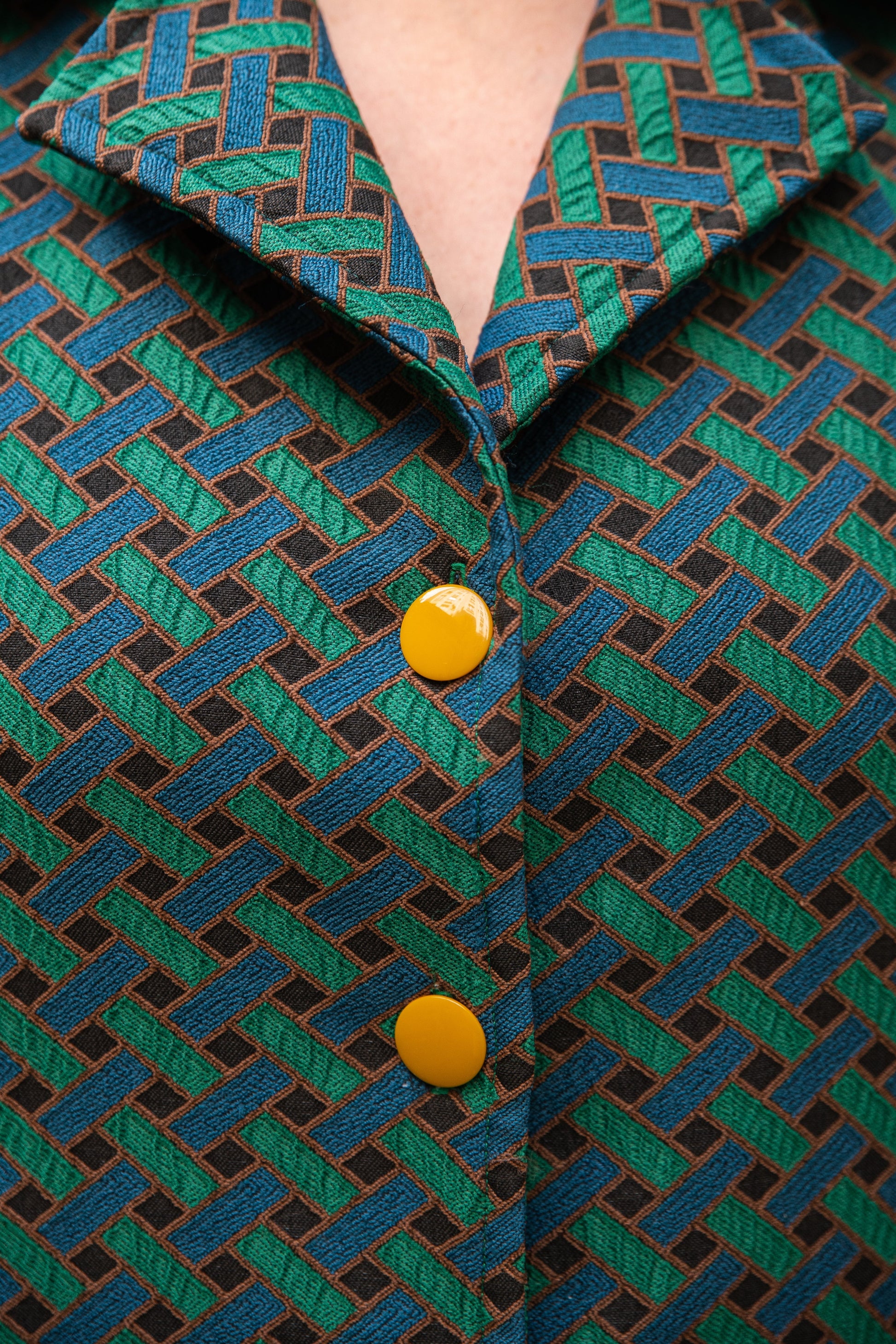 Gros plan sur le col chemisier à larges revers et les boutons ronds jaune moutarde de la robe jacquard de coton italien Carlis — tissage géométrique texturé vert forêt, bleu canard et noir, encolure V, boutonnage contrasté. Détail couture artisanale, robe élégante femme made in France.
