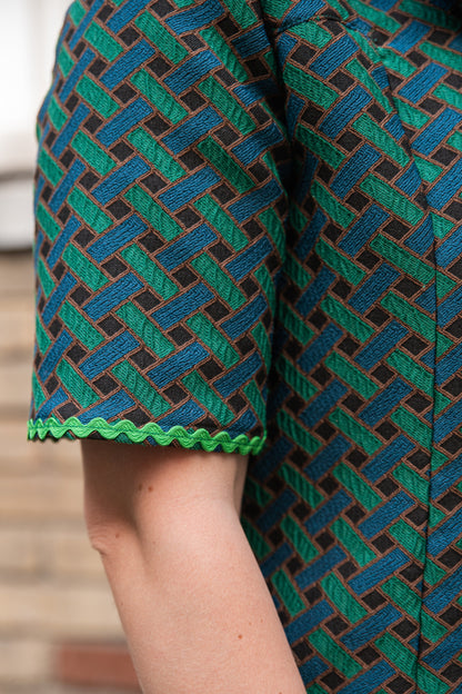 Gros plan sur la manche courte de la robe jacquard de coton italien Carlis — tissage géométrique texturé vert forêt, bleu canard et noir, finition passepoil ric-rac vert vif en bas de manche. Détail couture artisanale, robe élégante femme made in France.
